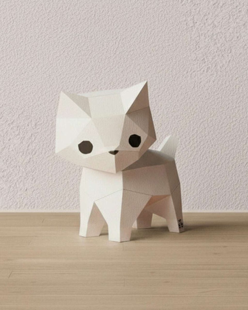 Chat blanc en papercraft 3D Moki Moki, figurine de style origami en papier à monter soi-même, posé sur un sol en bois clair devant un mur texturé.