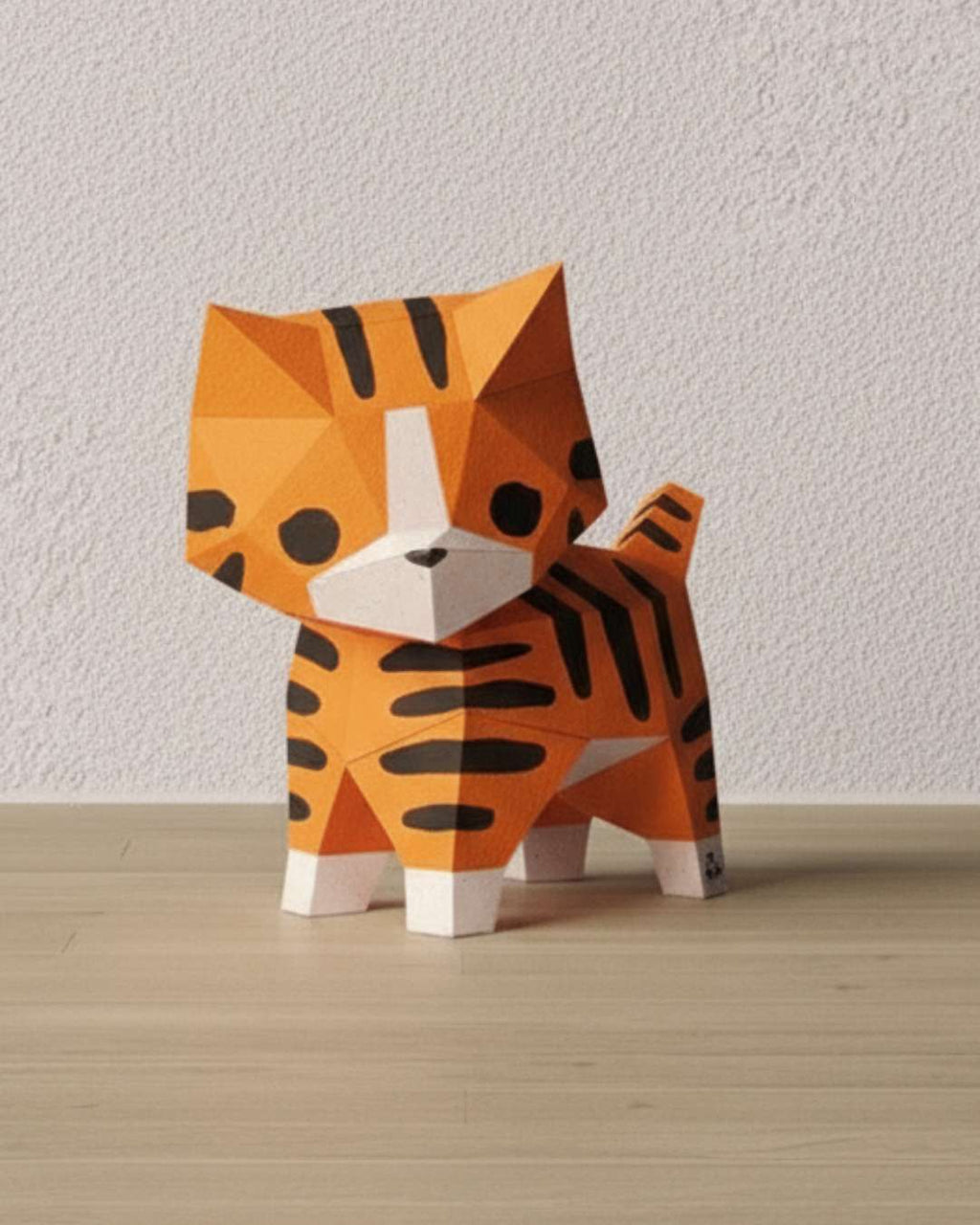 Chat roux en papercraft 3D Moki Moki de style origami, figurine en papier à monter soi-même, posée sur un sol en bois clair devant un mur texturé.