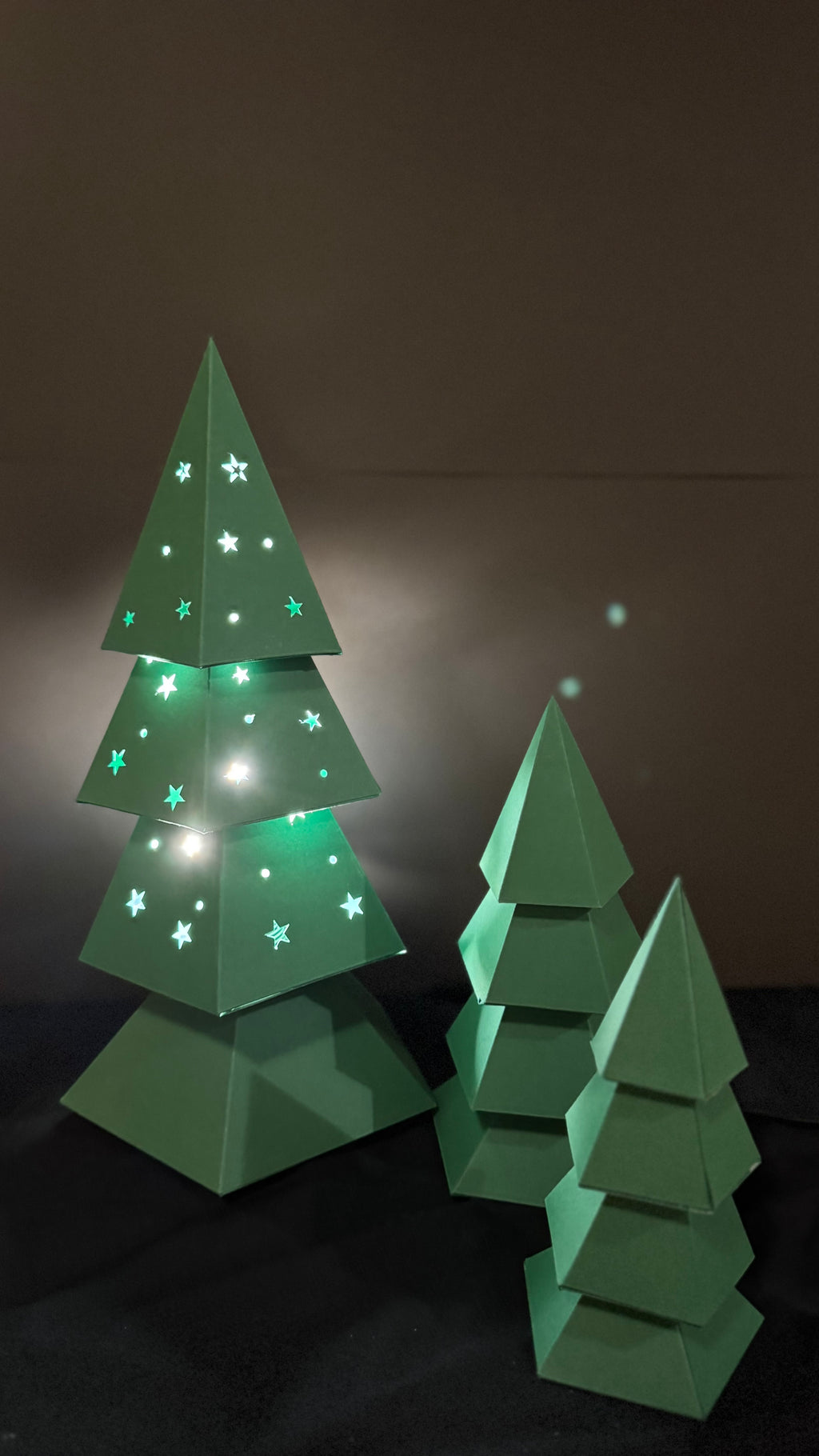 Sapin de noël lumineux en papercraft 3D avec découpes étoilées, entouré de sapins décoratifs en papier vert, création Moki Moki.
