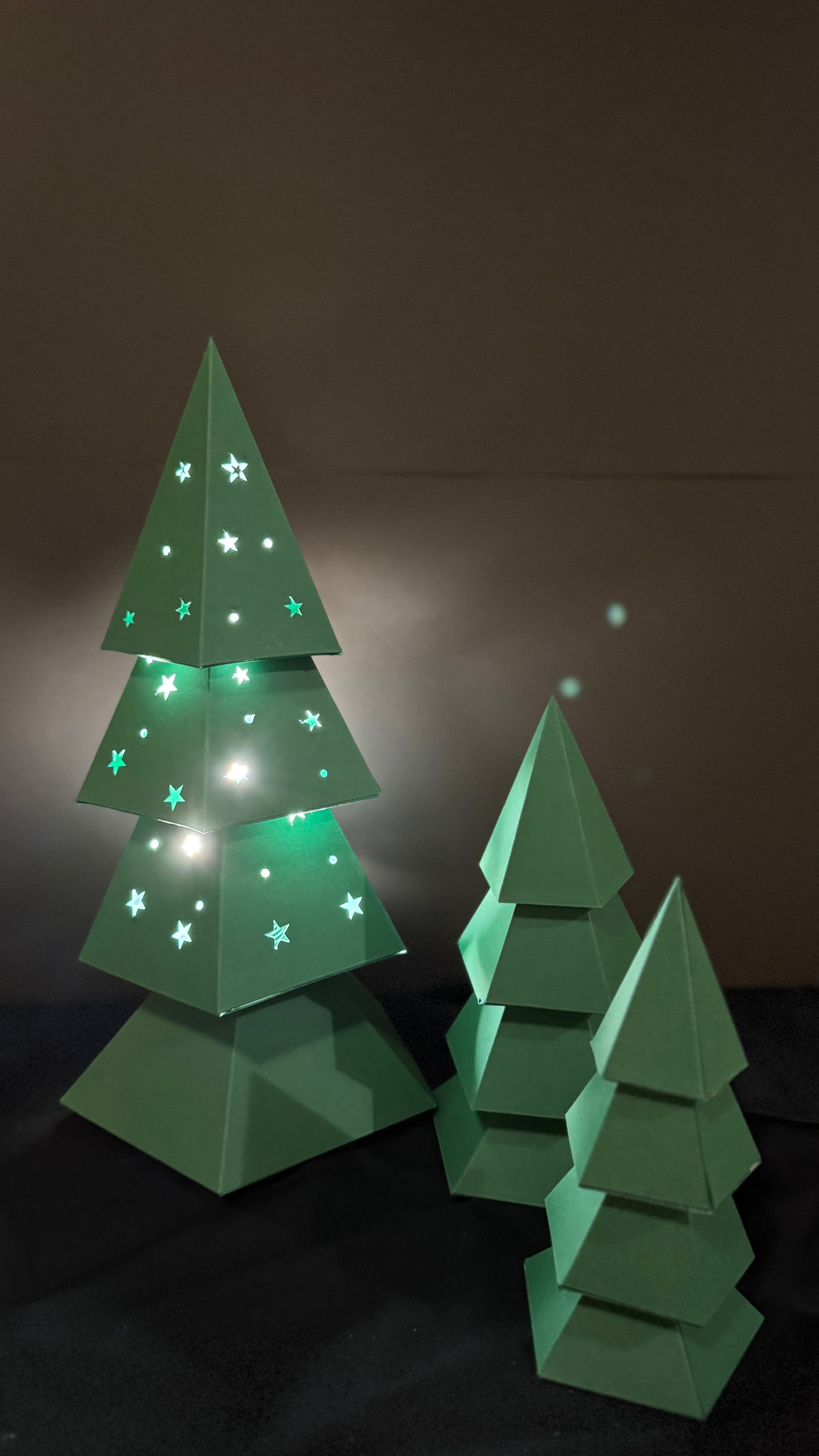 Sapin de noël lumineux en papercraft 3D avec découpes étoilées, entouré de sapins décoratifs en papier vert, création Moki Moki.