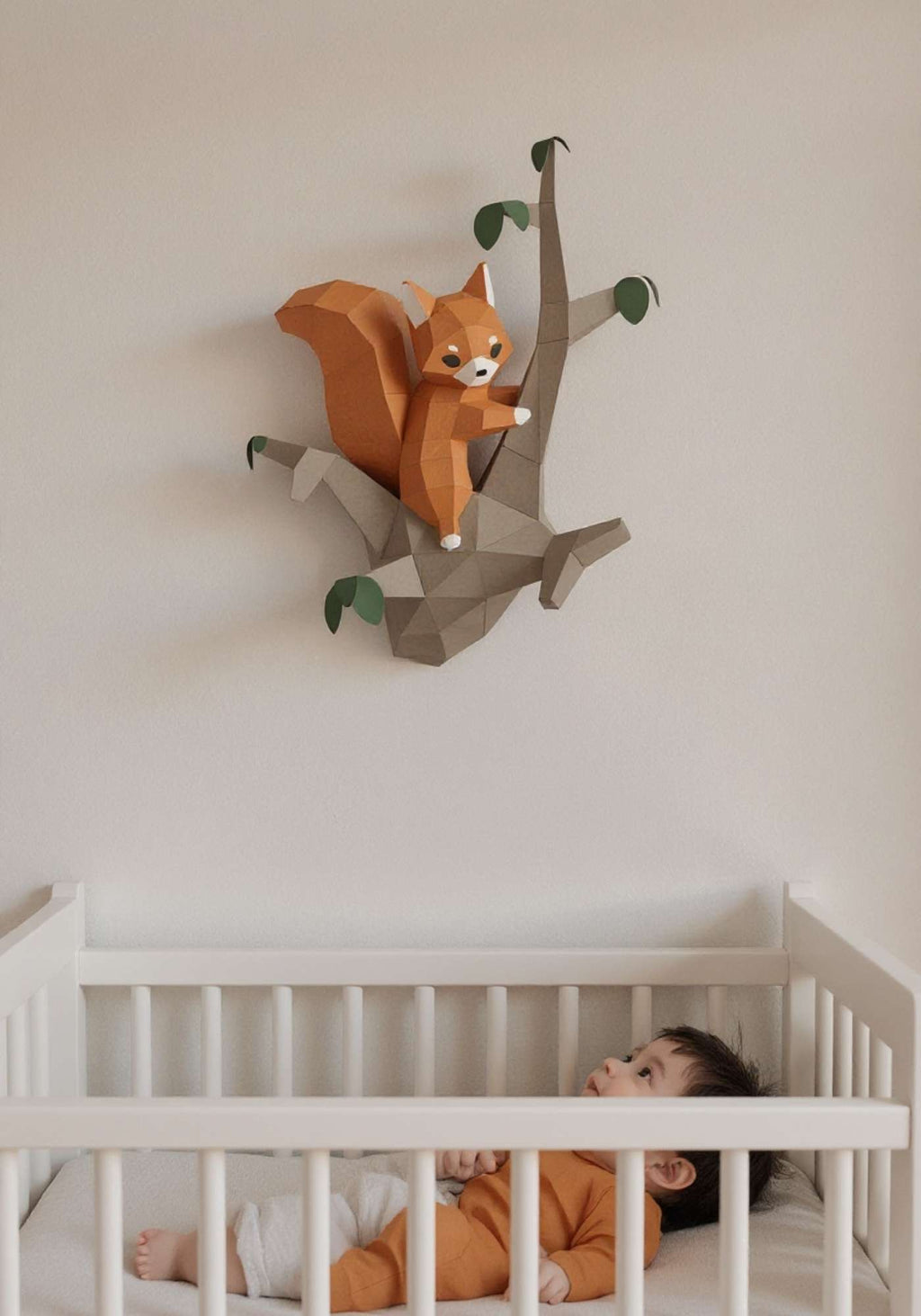 Wall art écureuil pour enfant en papier style origami 3D. Créée en papercraft 3D, l'applique murale est un écureuil sur la branche d'un arbre. Sur la photo, l'animal de la forêt est accroché sur le mur d'une chambre d'enfant. On voit un bébé dans son lit en train de regarder l'écureuil qui est accroché au-dessus du lit.