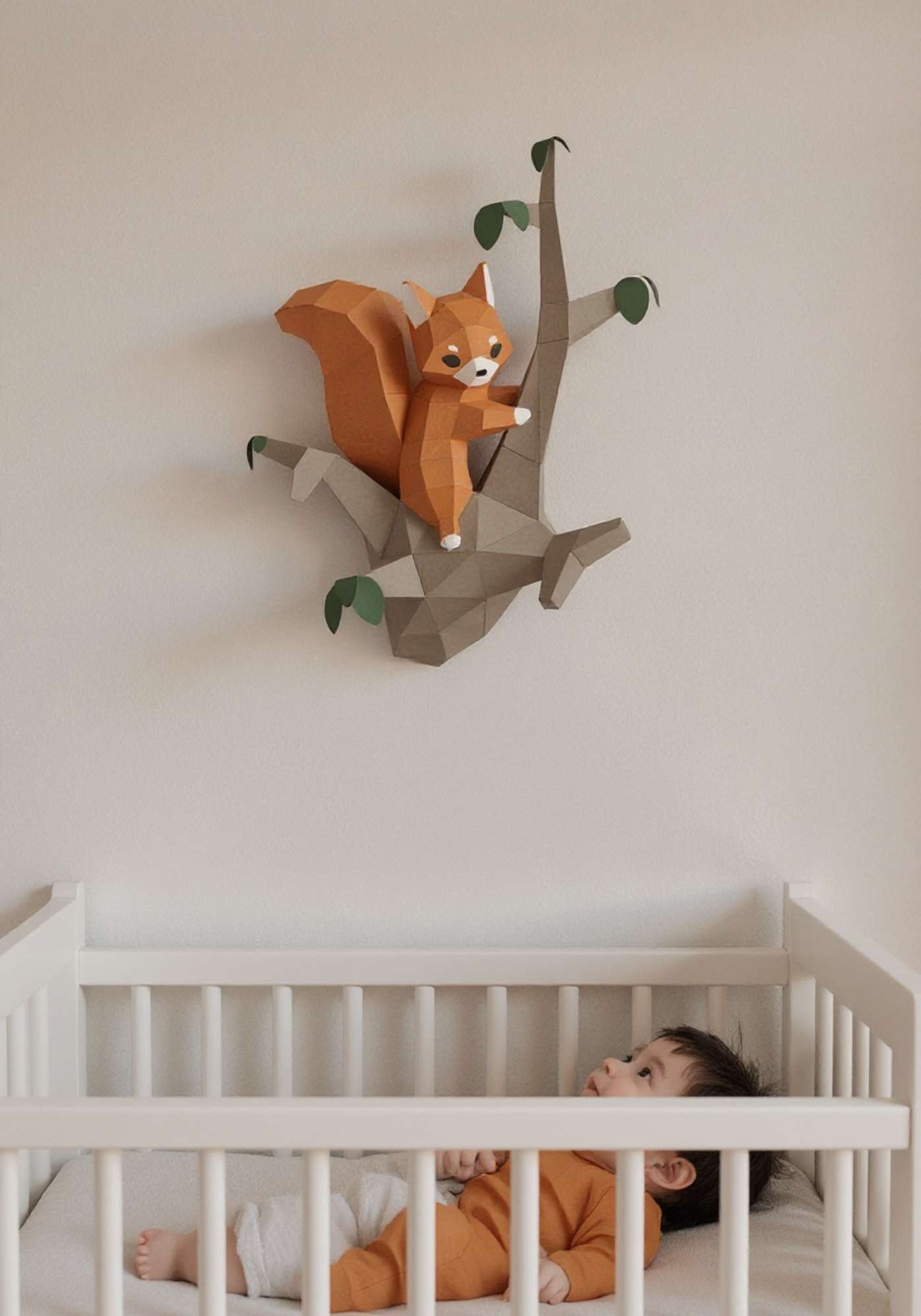 Wall art écureuil pour enfant en papier style origami 3D. Créée en papercraft 3D, l'applique murale est un écureuil sur la branche d'un arbre. Sur la photo, l'animal de la forêt est accroché sur le mur d'une chambre d'enfant. On voit un bébé dans son lit en train de regarder l'écureuil qui est accroché au-dessus du lit.