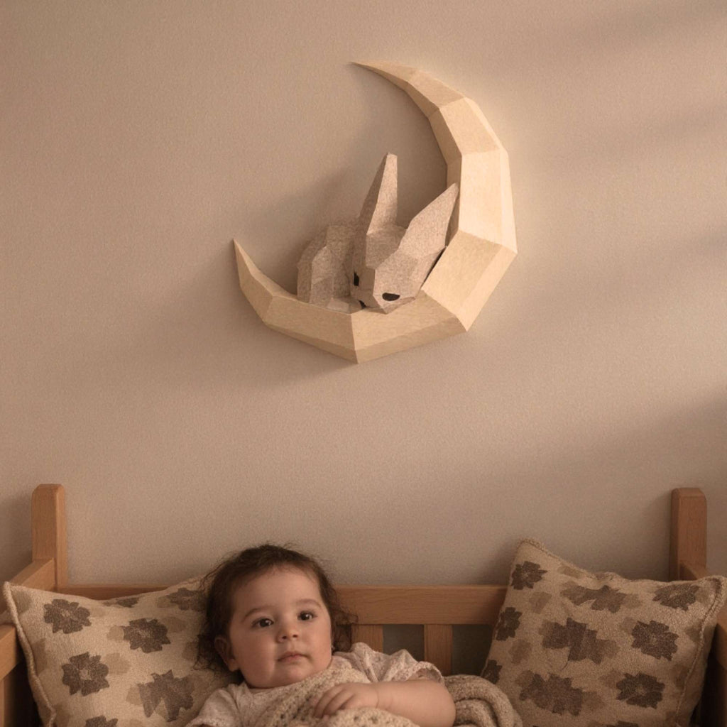 Wall art lapin pour enfant en papier style origami 3D. Créée en papercraft 3D, l'applique murale est un petit lapin en train de dormir sur sa lune. L'applique est accrochée sur le mur d'une chambre d'enfant. On voit un bébé en train de s'endormir avec le wall art lapin accroché au-dessus du lit.