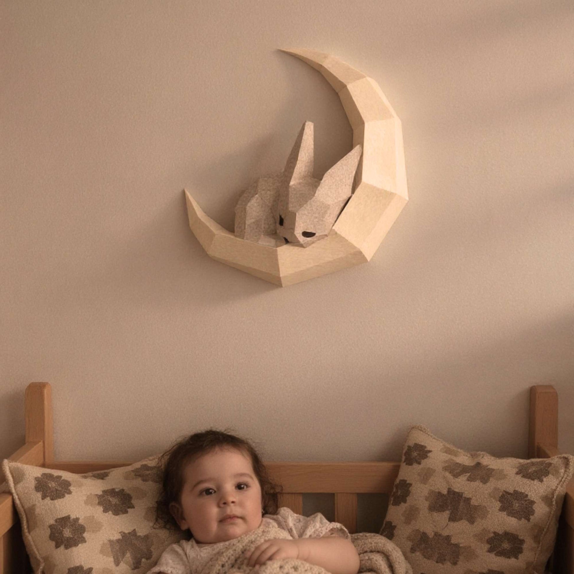 Wall art lapin pour enfant en papier style origami 3D. Créée en papercraft 3D, l'applique murale est un petit lapin en train de dormir sur sa lune. L'applique est accrochée sur le mur d'une chambre d'enfant. On voit un bébé en train de s'endormir avec le wall art lapin accroché au-dessus du lit.
