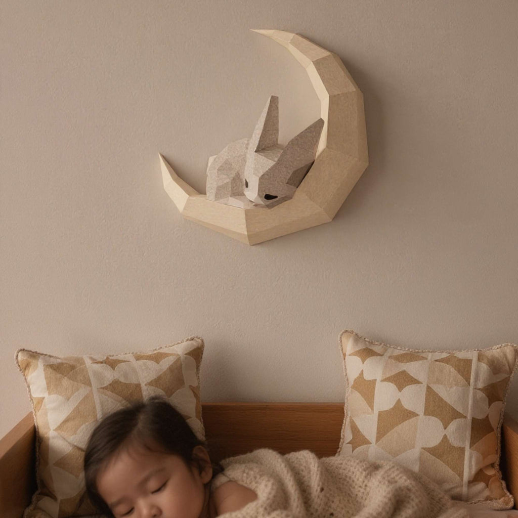 Applique murale lapin pour enfant en papier style origami 3D. Le wall art est un petit lapin en train de dormir sur une lune. L'applique est accrochée sur le mur d'une chambre de bébé. On voit un bébé dormant avec le wall art lapin accroché au-dessus du lit.