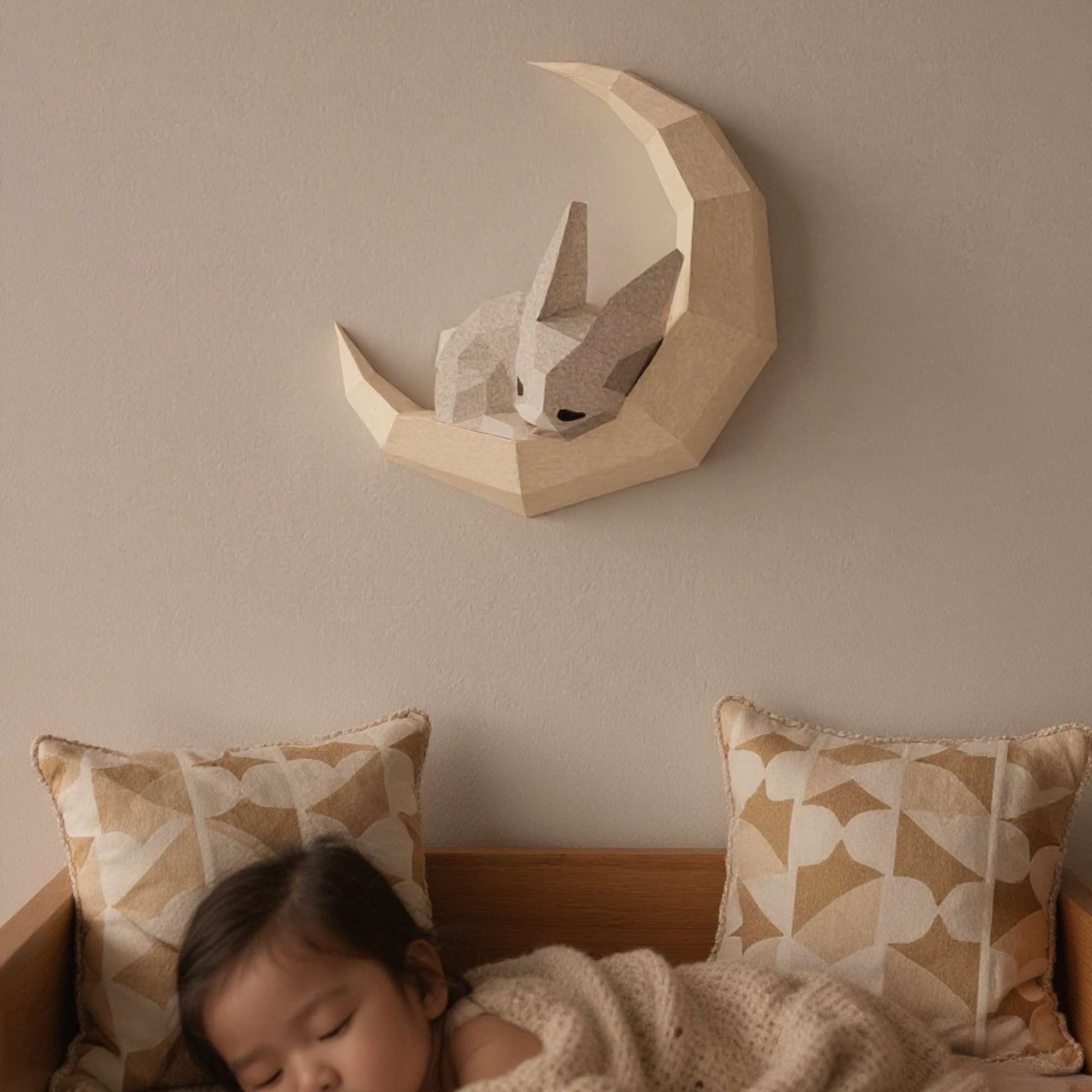 Applique murale lapin pour enfant en papier style origami 3D. Le wall art est un petit lapin en train de dormir sur une lune. L'applique est accrochée sur le mur d'une chambre de bébé. On voit un bébé dormant avec le wall art lapin accroché au-dessus du lit.