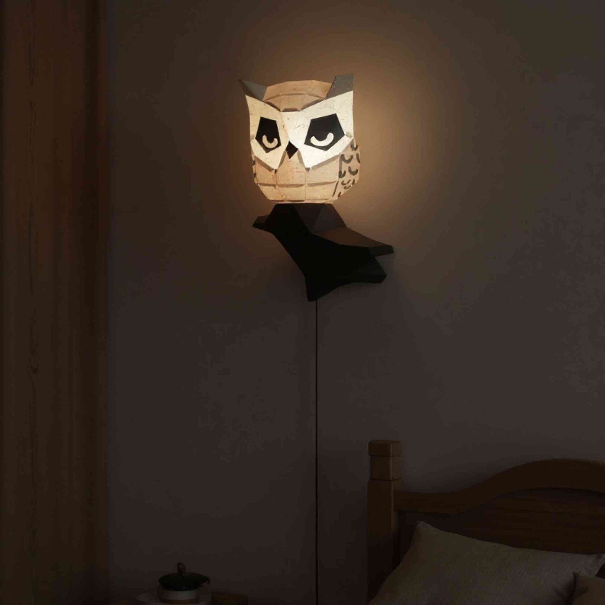La veilleuse applique murale hibou en papier style origami 3D est perché sur sa branche dans une chambre de petit enfant. La veilleuse est allumée et révèle la couleur naturelle du hibou. L'éclairage de ce wall art est doux et parfait pour des nuits tranquilles et apaisées.