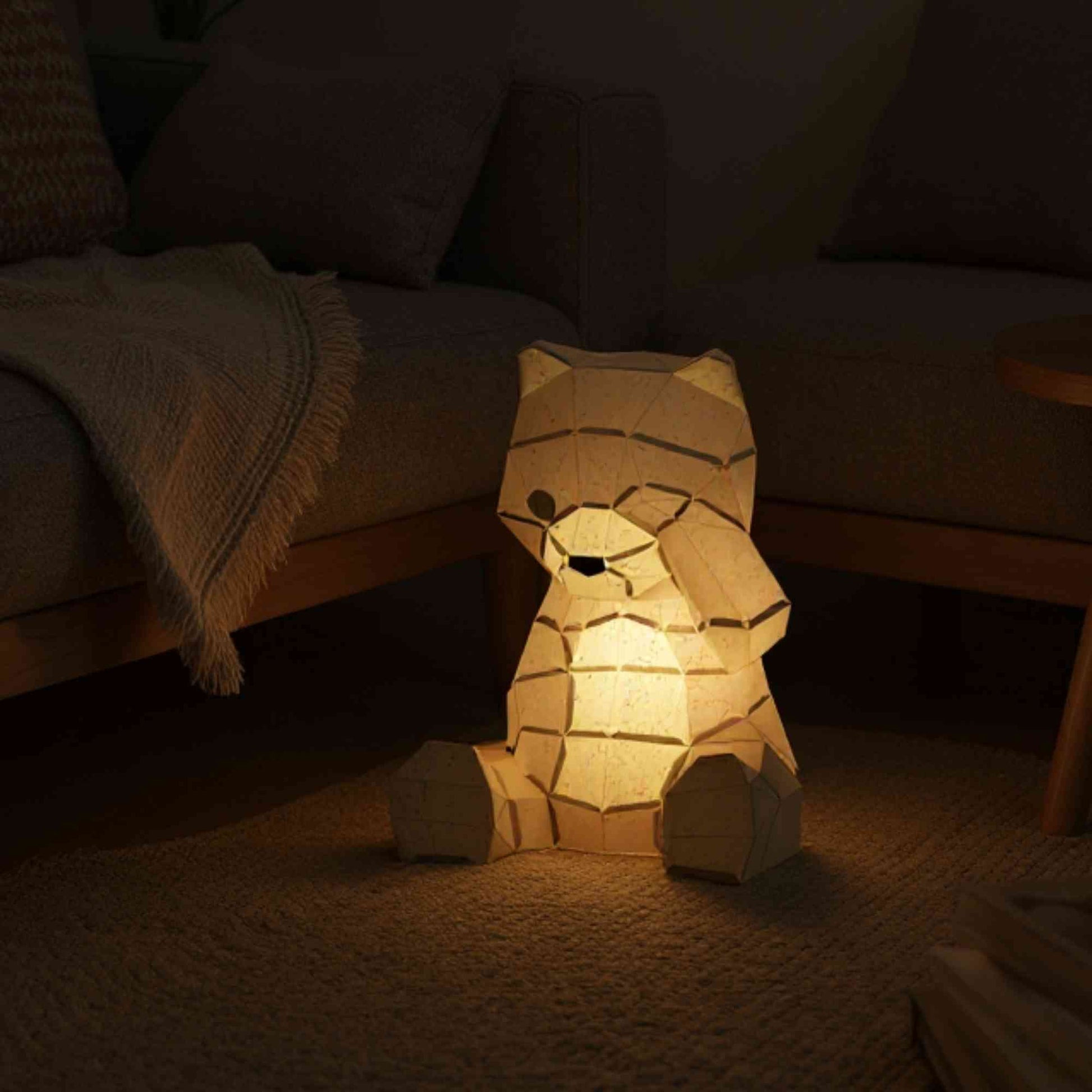 La lampe ourson en papier style origami 3D est dans un salon en décoration d'intérieur ou dans une chambre de bébé. La veilleuse est allumée et révèle la couleur naturelle de l'ours.