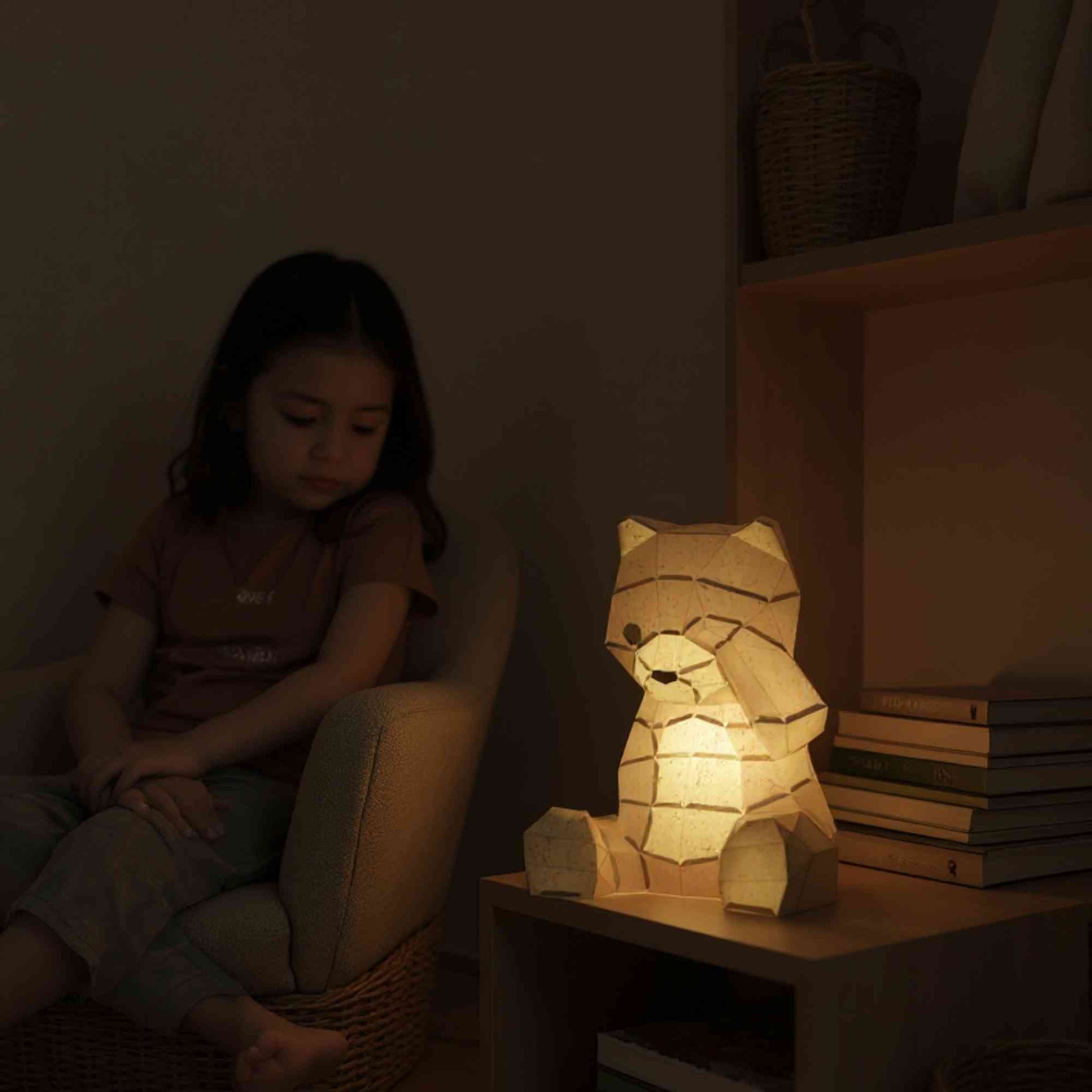 La veilleuse bébé et enfant ourson en papier style origami 3D est sur une table de chevet dans une chambre de petite fille. La veilleuse est allumée et révèle la couleur naturelle de l'ours. Une petite fille est assise à côté en train de le regarder. La lumière est douce.