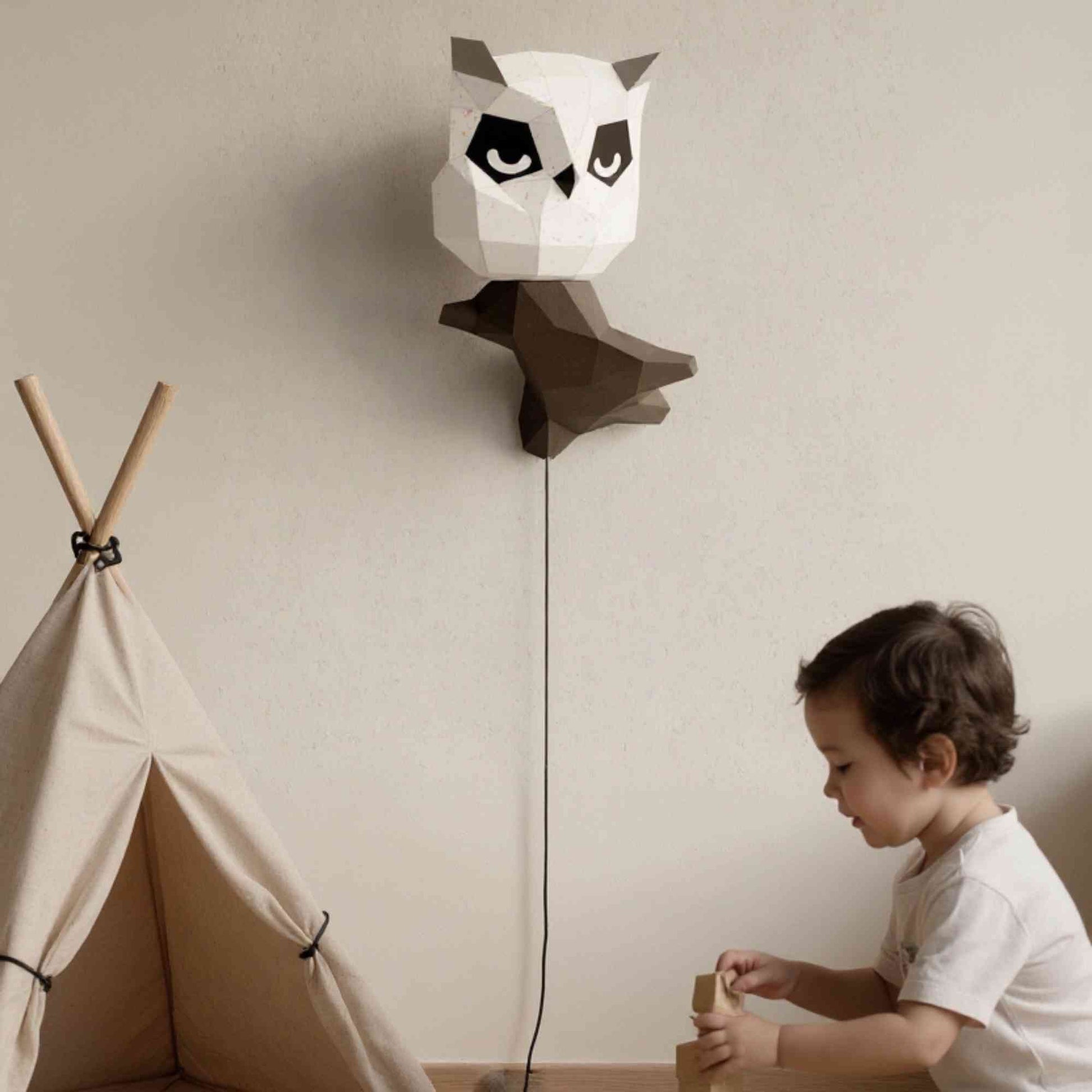 L'applique murale wall art hibou en papier style origami 3D est perché sur sa branche dans une chambre de petit garçon ou petite fille. La lampe veilleuse est éteinte et révèle le papier créatif blanc avec des dorures. On voit un petit enfant en train de jouer sous le hibou.
