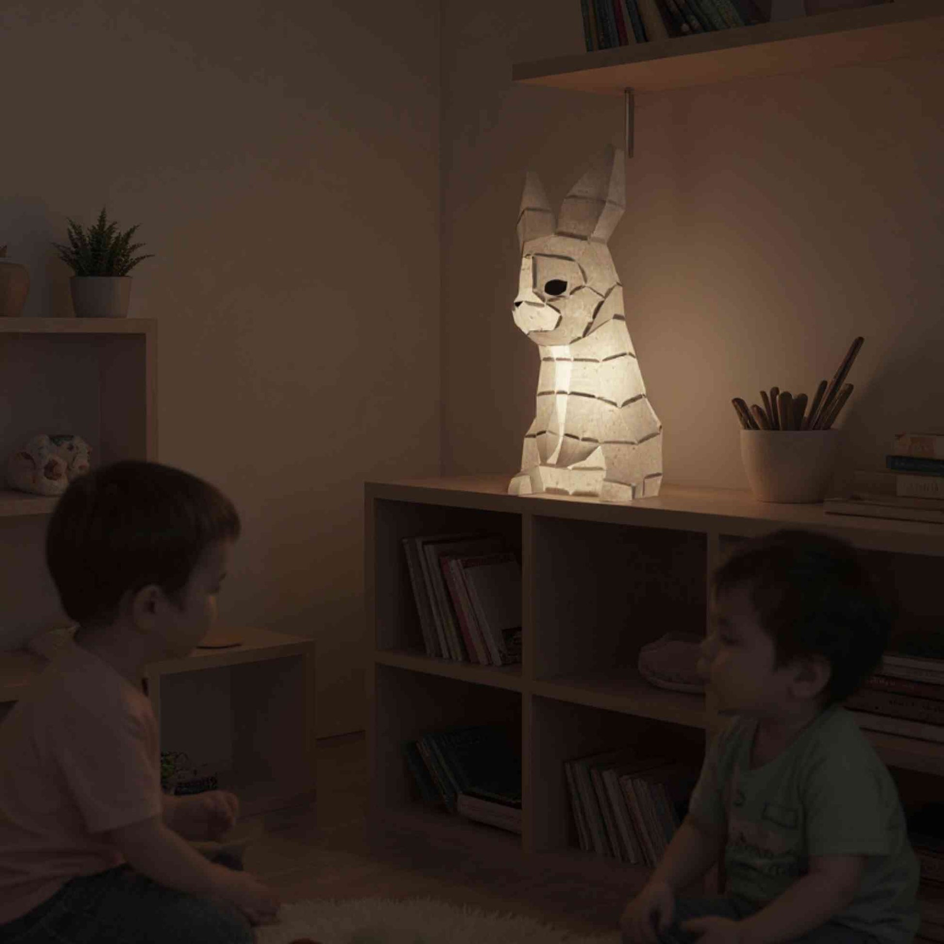 La veilleuse bébé et enfant lapin en papier style origami 3D est sur une commode dans une chambre d'un petit garçon. Deux enfants sont en train de jouer sous la lumière douce et naturelle de la lampe lapin.