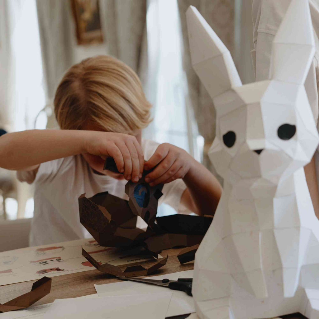 Enfant en train de monter un animal en papier en origami 3D. En premier plan, on voit qu'il a déjà construit le lapin veilleuse.