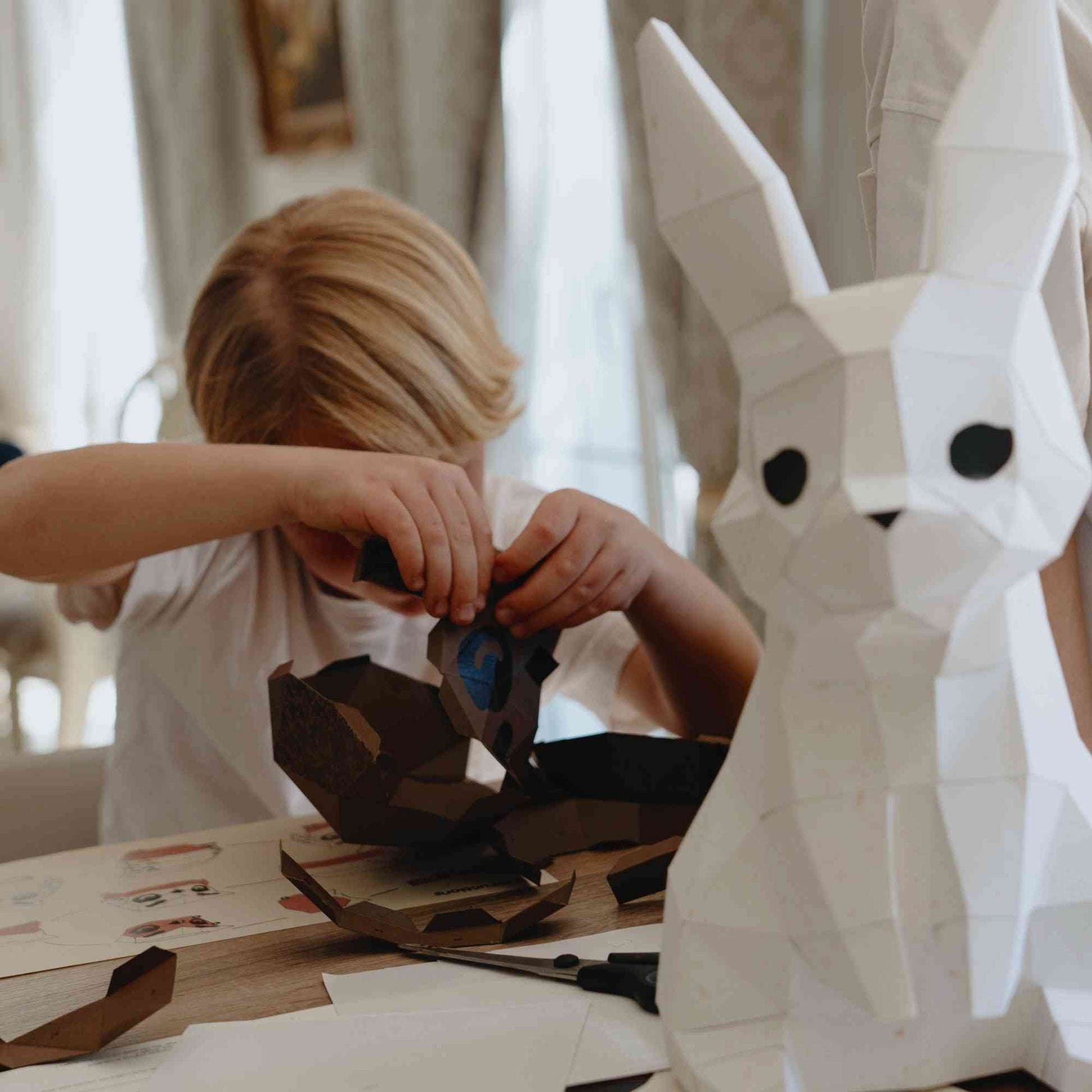 Enfant en train de monter un animal en papier en origami 3D. En premier plan, on voit qu'il a déjà construit le lapin veilleuse.
