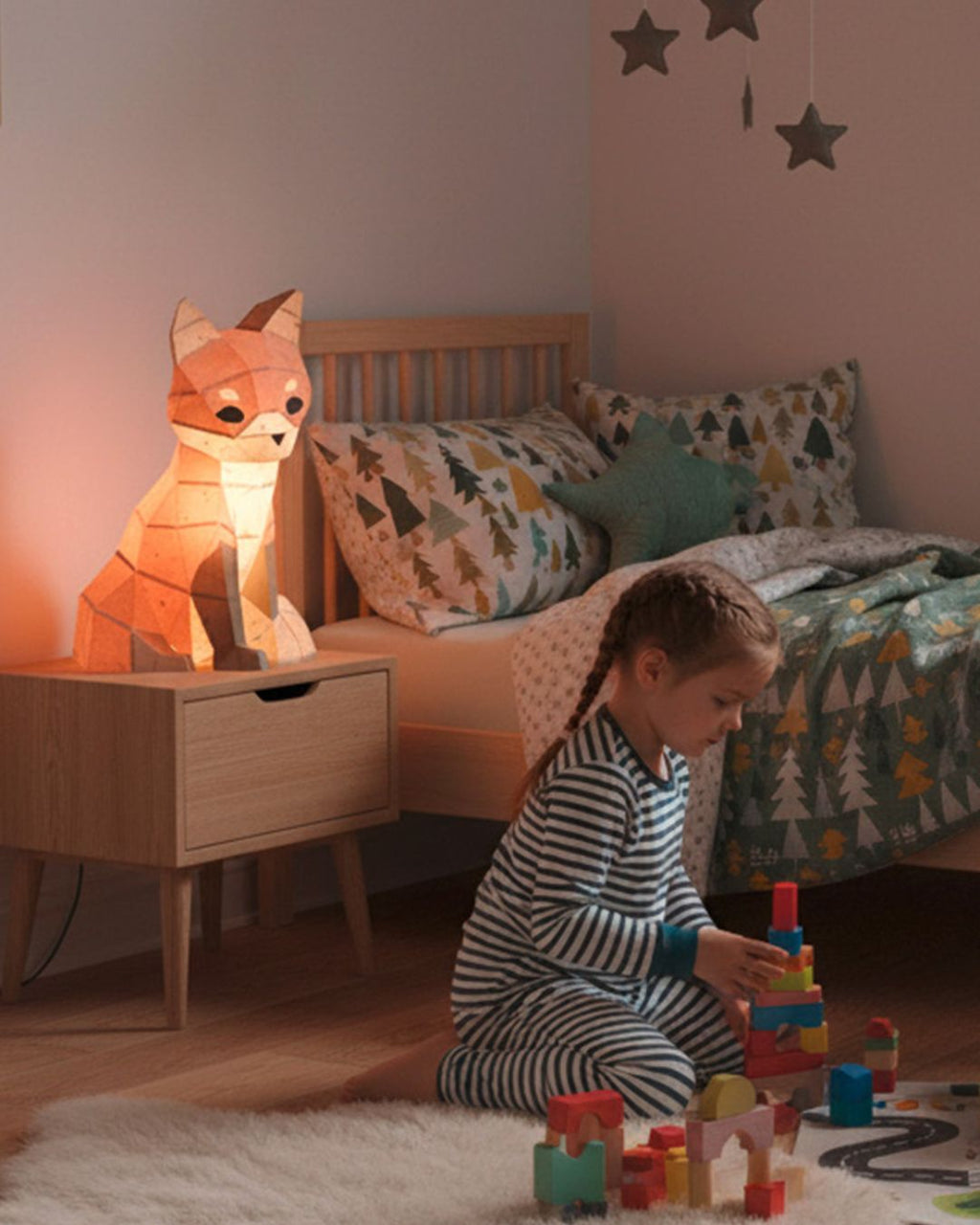 Lampe veilleuse renard en papercraft 3D de style origami, allumée sur une table de nuit dans une chambre d’enfant, petite fille jouant au sol dans une ambiance douce.