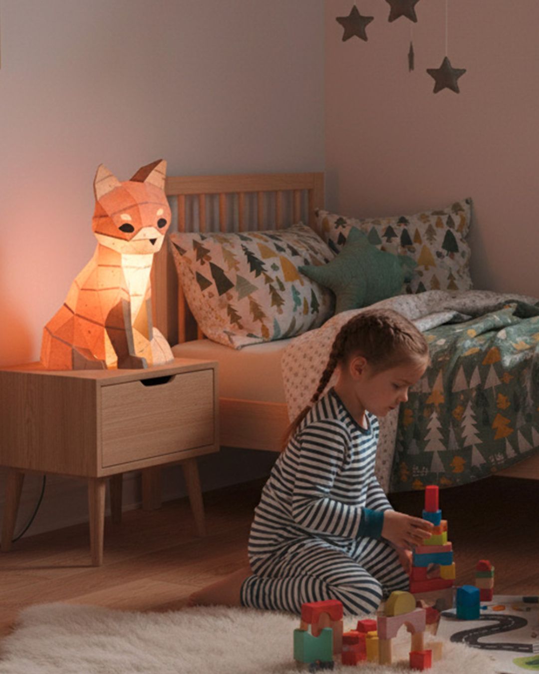 Lampe veilleuse renard en papercraft 3D de style origami, allumée sur une table de nuit dans une chambre d’enfant, petite fille jouant au sol dans une ambiance douce.